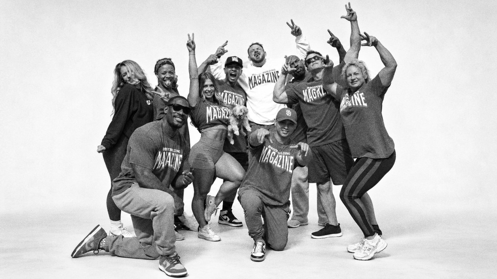 The AJ Bodybuilding Magazine team posing together in AJ Magazine shirts against a studio backdrop